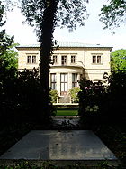 A grey slab, level with the ground framed by bushes and in the shade of the tree. In the background a fountain and a large two-storied house with a balcony.