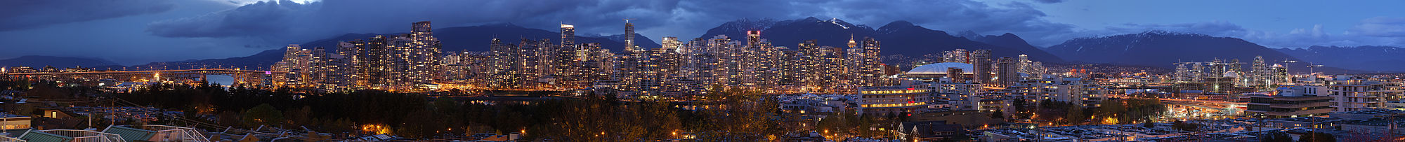 High-resolution panorama of a large, brightly-lit skyline at night. A mountain range lies in the background, and a bridge is visible on the left-hand side of the panorama.