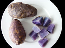 Two dark-skinned potatoes on a white plate. A further potato is cut into sections to show the variety's purple-blue flesh, placed at lower-right on the plate.