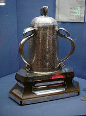 In a blue display cabinet is a bell shaped silver cup which is heavily engraved with patterns; it is adorned on its lid with a small silver elephant and both its large handles are silver cobras.